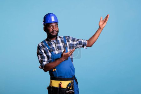 Foto de Construction worker raises hands to show an imaginary product, making advertising presentation with empty hands. Showing something as advertising, presenting gesture on blue background in studio. - Imagen libre de derechos