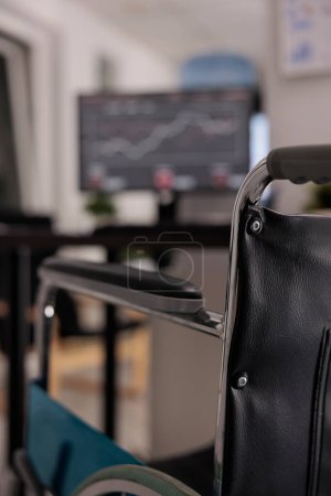 Photo for Close up view of wheelchair for employee with walking disability in an empty modern workspace. Desktop computer with graphs and annual statistics, wheelchair accessible office. - Royalty Free Image