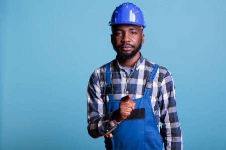 Téléchargez les photos : African american man in coveralls pointing forward, looking at camera making the choose you gesture on blue background. Confident builder pointing with index finger in studio shot. - en image libre de droit
