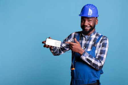 Photo for African american construction worker in protective helmet pointing finger at cell phone with empty screen isolated on blue background. Builder in studio shot holding advertising blank display. - Royalty Free Image