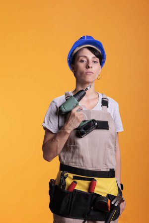 Photo for Female industrial builder holding power drill gun in front of camera, using electric drilling tool before renovation project. Construction worker wearing blue helmet and building uniform. - Royalty Free Image