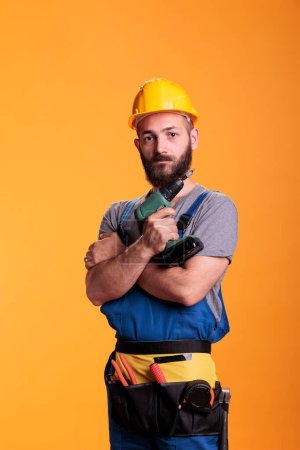 Téléchargez les photos : Engineering repairman holding cordless electric drill, posing in studio. Male construction worker using power drilling gun to screw nails, wearing overalls and and hardhat on camera. - en image libre de droit