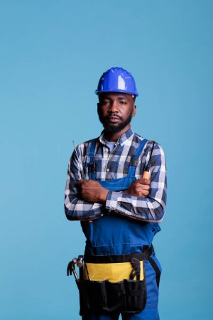 Téléchargez les photos : Portrait of construction professional in front of camera holding screwdriver in studio shot. African american builder wearing tool belt and work uniform isolated on blue background. - en image libre de droit