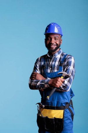 Téléchargez les photos : Happy African american builder holding hammer posing in studio against blue background. Workman in hard hat and construction overalls, safety uniform in construction industry. - en image libre de droit