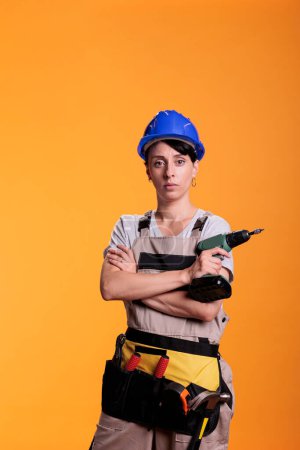 Photo for Professional handywoman holding electric power drill, wearing hardhat and overalls to renovate walls. Serious construction worker using nail drilling gun, working tool on studio background. - Royalty Free Image