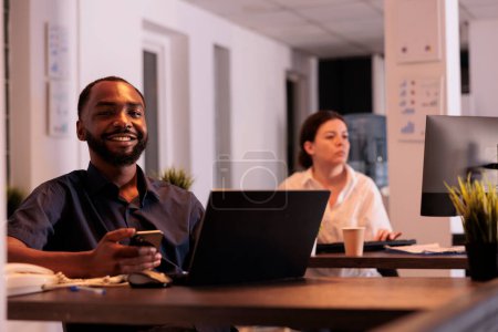 Téléchargez les photos : Smiling african american man working on report on laptop, using smartphone software in coworking space portrait. Happy project manager looking at camera at workplace desk - en image libre de droit