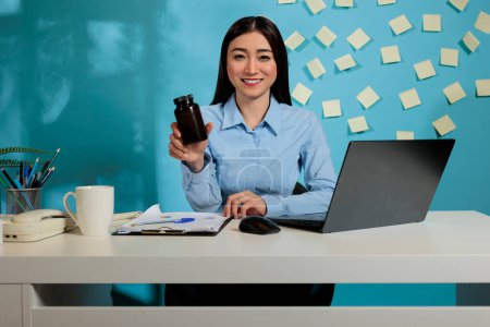 Téléchargez les photos : Asian professional woman sitting at modern office desk holding black bottle of pills. Corporate employee at work station with dark container of tablets looking at camera. - en image libre de droit