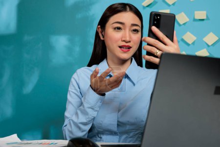 Téléchargez les photos : Asian businesswoman holding a virtual meeting with a smartphone and wireless headset sitting at her desk. Concentrated employee attending a remote conference with a mobile phone. - en image libre de droit