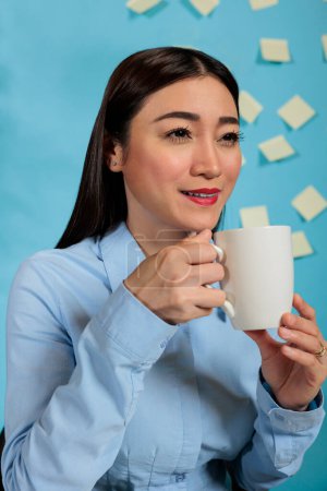 Foto de Asian woman taking a break from work using laptop drinking coffee from cup in modern space. Casual startup employee working with statistics in a business workplace, close up. - Imagen libre de derechos