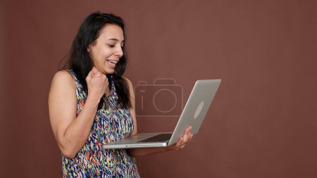 Téléchargez les photos : Modèle féminin heureux tenant ordinateur portable sur la caméra, en utilisant le site de médias sociaux pour naviguer sur Internet et discuter. Se sentir satisfait de gadget PC portable sans fil avec réseau en ligne. - en image libre de droit