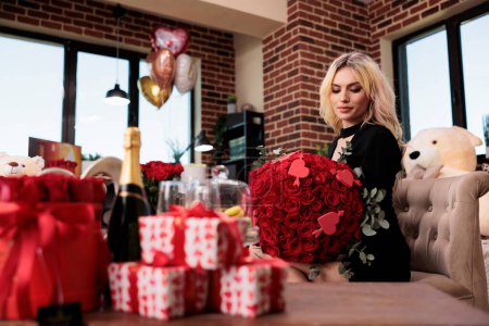 Foto de Mujer sosteniendo ramo de rosas rojas con expresión facial tranquila en la sala de estar llena de regalos de San Valentín. Saludo romántico aniversario, chica rubia sentada con flores, plano mediano - Imagen libre de derechos