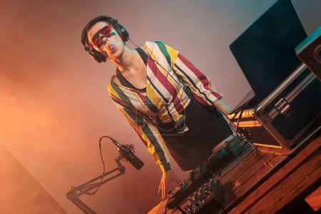 Téléchargez les photos : Flirty DJ femme agissant stupide avec la langue sur, s'amuser dans le studio avec des lumières. Musicien funky ludique mêlant musique techno avec mixeur ou platines tournantes, confiant et décontracté. - en image libre de droit