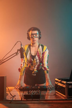 Photo for Female DJ standing at turntables and mixing equipment, using audio instruments or mixer to perform techno music. Serious musician with crazy make up doing remix with bass key and buttons. - Royalty Free Image