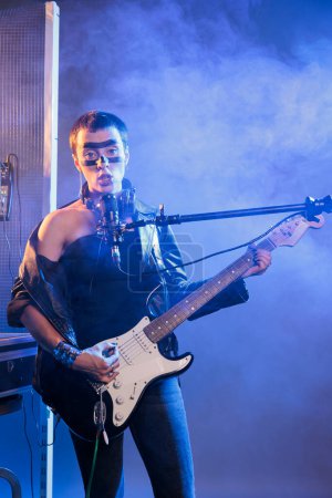 Foto de Punk rocker with cool make up singing alternative rock, playing guitar and doing concert show in studio. Ecstatic talented guitarist performing heavy metal music live with microphone and instrument. - Imagen libre de derechos