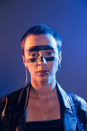 Téléchargez les photos : Female musician with crazy make up black lines wearing leather jacket and preparing to do live rock performance with heavy metal music. Funky artist posing in studio with dark lights and smoke. - en image libre de droit