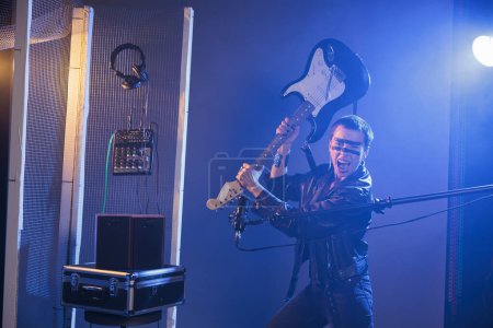 Téléchargez les photos : Cool stylish musician holding bass guitar to smash and act crazy, performing punk rock music and screaming loud in studio. Rocker with leather jacket fooling around and throwing musical instrument. - en image libre de droit