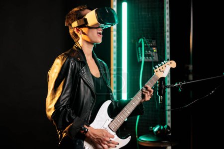 Photo for Woman musician playing at electric guitar in sound studio while wearing virtual reality headset for concert simulation, enjoying music performance. Rebel performer working at new heavy metal album - Royalty Free Image