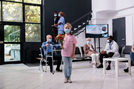 Photo for Child looking at camera while holding bouquet of flowers waiting for grandfather to finish examination with doctor. People wearing medical face mask to prevent infection with covid19 in hospital lobby - Royalty Free Image