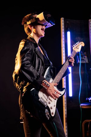 Téléchargez les photos : Rebel performer playing heavy metal song using electric guitar while wearing vr headset to have rock concert simulation, enjoying experience in music studio. Woman musician working at new grunge album - en image libre de droit