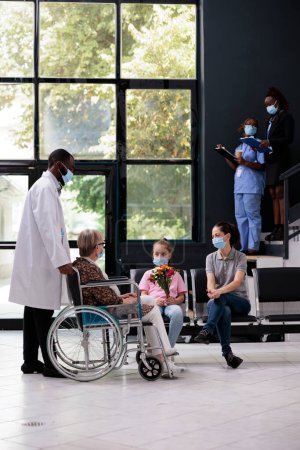 Photo for African american medic bringing disabled senior woman to family after medical consultation during checkup visit examination. Little girl holding bouquet flowers for grandmother. Virus pandemic - Royalty Free Image