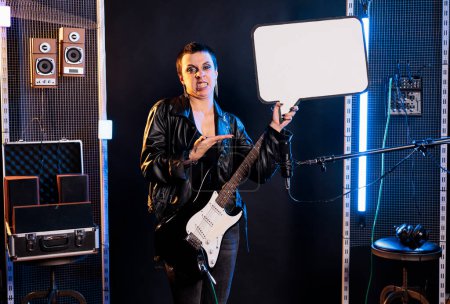 Téléchargez les photos : Woman musician holding blank white cardboard for advertise text while wearing electric guitar preparing to play rock song in studio. Guitarist using electric instrument to perform heavy metal music - en image libre de droit