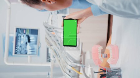 Photo for Vertical video: Dentist looking at mobile phone with green screen in dentistry office. Woman working with isolated background and mockup template on smartphone for stomatological care - Royalty Free Image