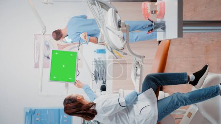 Téléchargez les photos : Vidéo verticale : Le dentiste et l'infirmière analysent la radiographie dentaire et parlent de contrôle dentaire, travaillant avec un écran vert horizontal sur l'écran. Spécialiste et homme regardant la radiographie et la clé chromatique - en image libre de droit