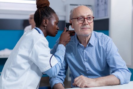 Photo for Clinic otology specialist consulting senior patient using otoscope to check ear infection. Hospital otologist examining sick retired man internal ear condition while in doctor cabinet. - Royalty Free Image