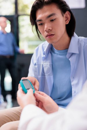 Photo for Elderly physician measuring insulin and glucose level with glucometer while doing health examination with diabetic asian patient. Using insturment to check blood sample in hospital waiting area - Royalty Free Image