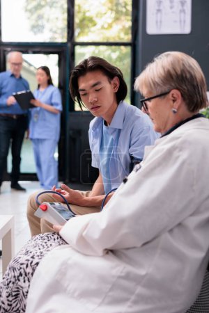 Photo for Elderly cardiologist measuring asian patient hypertension and blood pressure to do heart exam with tonometer in hospital waiting area. Doctor consulting young adult during checkup visit consultation - Royalty Free Image