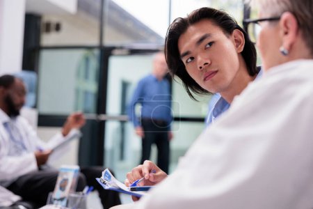 Photo for Senior physician explaining health care treatment to asian man during checkup visit appointment in hospital waiting room. Young patient holding clipboard filling medical report before examination - Royalty Free Image