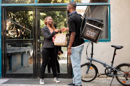 Téléchargez les photos : Office food delivery, courier giving customer order paper bag. Man holding restaurant takeaway meal, smiling employee taking lunch package near company building, wide shot - en image libre de droit