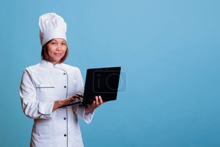Photo for Cheerful cook with apron holding laptop computer checking gastronomy recipe ingredient list, preparing culinary dish. Cheerful asian person working in food industry cooking healthy meal - Royalty Free Image