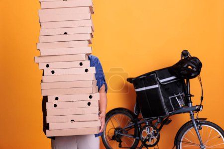 Foto de Large stack of pizza being carried by restaurant worker in blue uniform for one of restaurant clients during lunk time. Elderly asian employee deliverying food order with bike, take away concept - Imagen libre de derechos