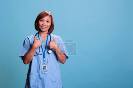 Téléchargez les photos : Médecin infirmier souriant portant un uniforme médical sur fond bleu montrant les pouces vers le haut geste en studio. Assistante âgée avec expression joyeuse faisant signe positif avec les mains, service de soins de santé - en image libre de droit