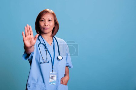 Téléchargez les photos : Asian medical assistant doing stop gesture with hands while working at illness expertise planning health care treatment for sick patient. Serious nurse standing in studio with blue background - en image libre de droit
