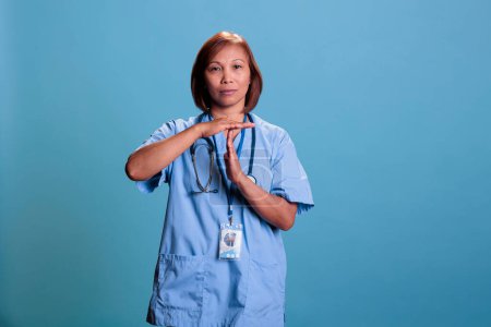 Téléchargez les photos : Assistante médicale asiatique faisant stop signe avec les mains tout en travaillant à l'expertise de la maladie la planification des soins de santé pour les patients malades. Infirmière sérieuse debout en studio avec fond bleu - en image libre de droit