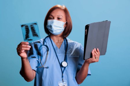 Téléchargez les photos : Elderly asian nurse analyzing lungs radiography checking medical expertise on tablet computer working at disease diagnosis for patient. Assistant wearing protective face mask against coronavirus - en image libre de droit