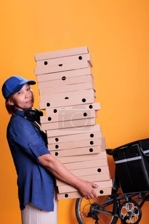 Téléchargez les photos : Pizzeria employee wearing blue t shirt and cap holding large stack of pizza delivering to client on bike. Food delivery worker standing in studio with yellow background. Food service and concept - en image libre de droit
