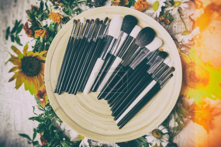 Foto de Diferentes tipos de pinceles de maquillaje en un plato junto a flores silvestres sobre fondo de madera - Imagen libre de derechos