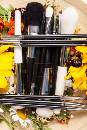 Téléchargez les photos : Different Make up brushes on plate next to wild flowers on wooden background - en image libre de droit