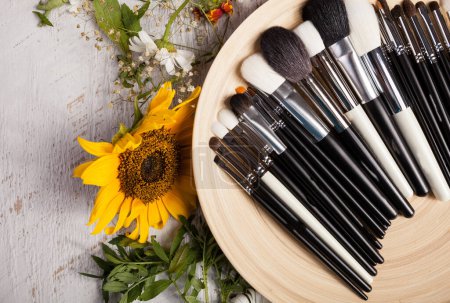 Téléchargez les photos : Different type of Make up brushes on a plate next to wild flowers on wooden background - en image libre de droit