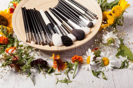 Téléchargez les photos : Professional Make up brushes on plate next to wild flowers on wooden background - en image libre de droit