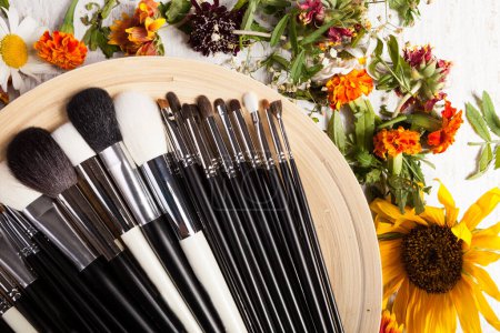 Foto de Diferentes tipos de pinceles de maquillaje en un plato junto a flores silvestres sobre fondo de madera - Imagen libre de derechos