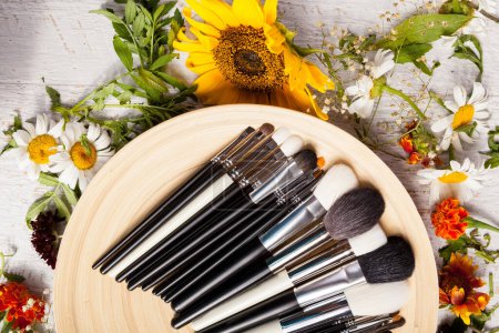 Photo for Different type of Make up brushes on a plate next to wild flowers on wooden background - Royalty Free Image