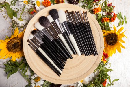 Téléchargez les photos : Différents types de pinceaux de maquillage sur une assiette à côté de fleurs sauvages sur fond en bois - en image libre de droit