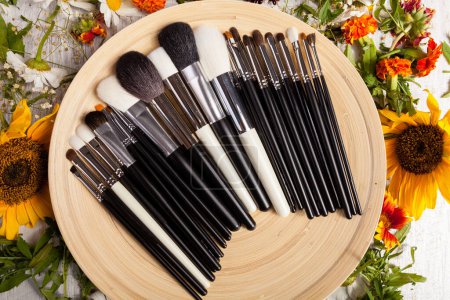 Téléchargez les photos : Différents types de pinceaux de maquillage sur une assiette à côté de fleurs sauvages sur fond en bois - en image libre de droit