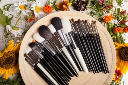 Téléchargez les photos : Différents pinceaux de maquillage sur plaque à côté de fleurs sauvages sur fond en bois - en image libre de droit