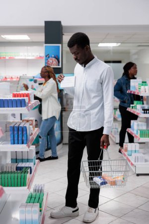 Foto de Hombre afroamericano de compras en la farmacia, la compra de medicamentos, instrucciones de lectura en el paquete de tabletas, de pie en el pasillo de la farmacia. Cliente elegir vitaminas, la compra de medicamentos, tiro ancho - Imagen libre de derechos