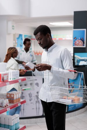 Photo for African american customer shopping in drugstore, buying supplements, reading instruction on tablets package. Young man choosing vitamins, purchasing medicaments in pharmacy store, medium shot - Royalty Free Image